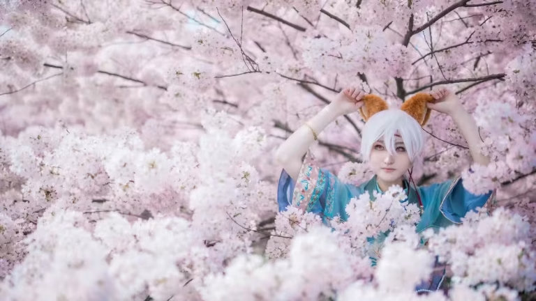 Professional Richmond cosplay photography capturing a white-haired fox spirit cosplayer adjusting her ears under the cherry blossoms at Larry Berg Flight Path Park, Metro Vancouver, British Columbia, Canada.