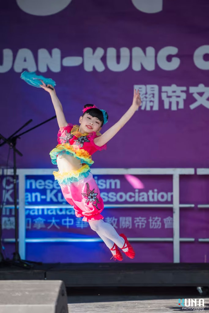 TJ Fest 2025 Kuna Photography: Vancouver Event Photographer. Young girl in pink & yellow costume performing a high jump during a traditional dance in BC.