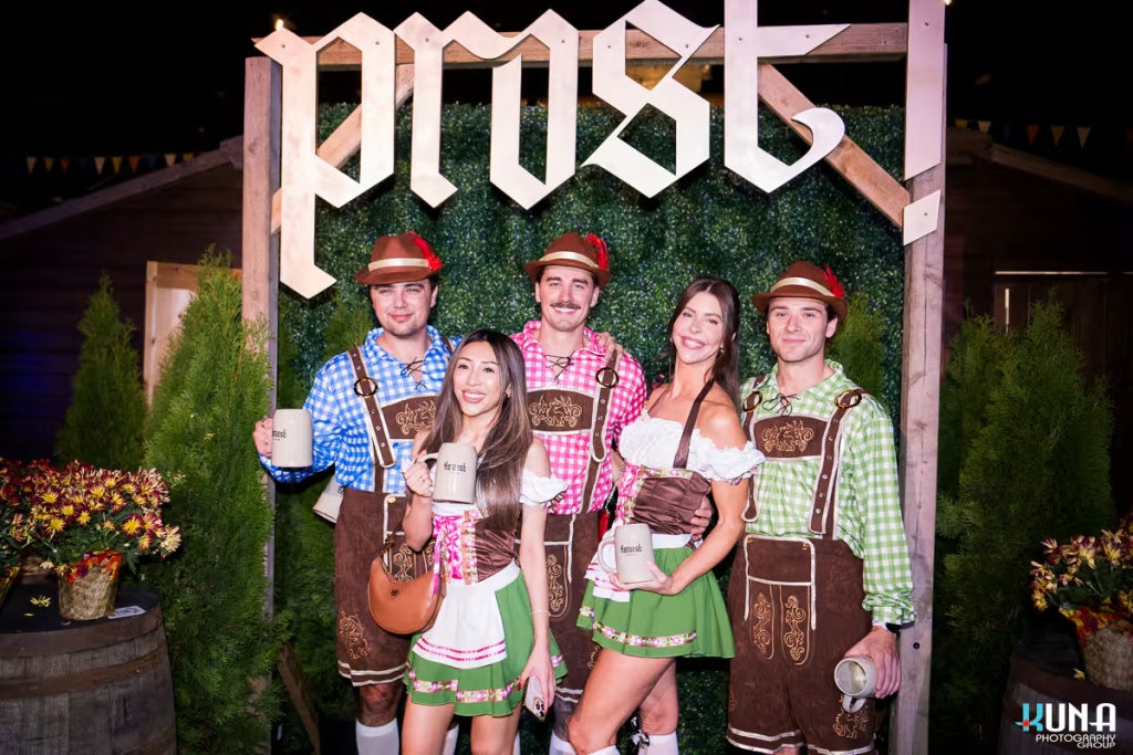vancouver event photography - A group of friends in colorful checkered lederhosen and dirndls posing under a "Prost!" sign at Harvest Haus Vancouver.