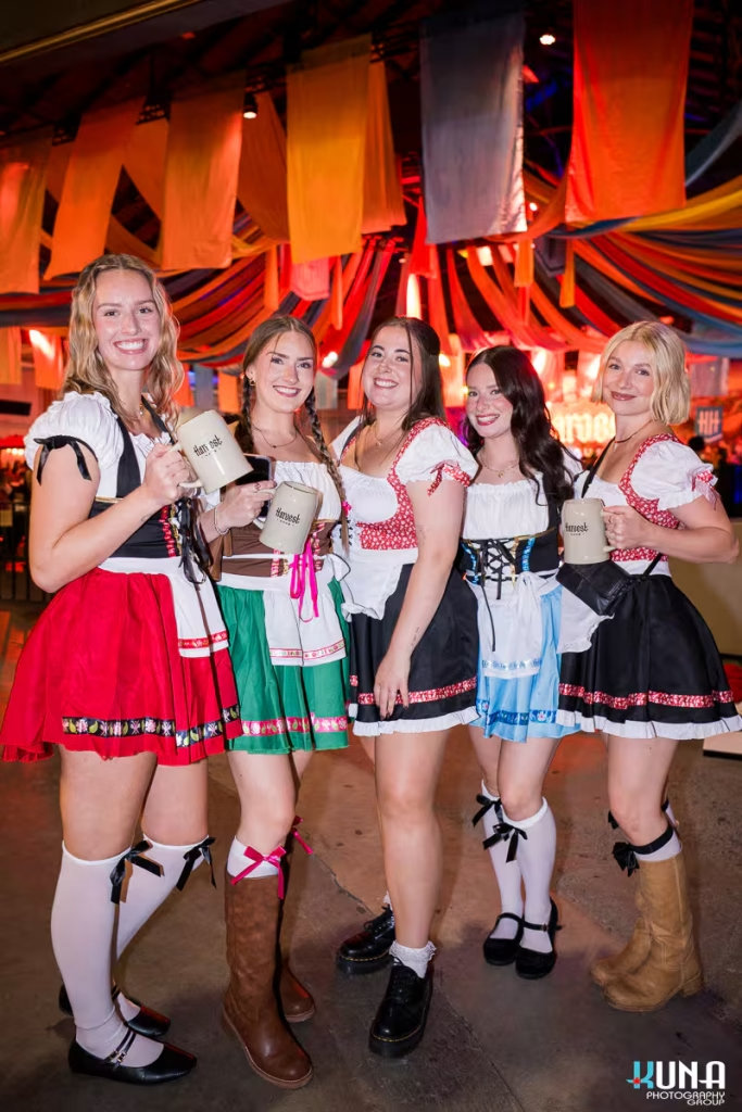 vancouver event photography - Five women in colorful dirndls and white knee-high socks posing together with beer steins under festive festival decorations.