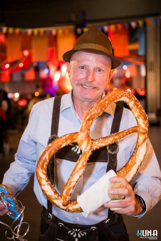 vancouver event photography - A man in a traditional Alpine hat and lederhosen smiling while holding a giant soft pretzel at Harvest Haus Vancouver.