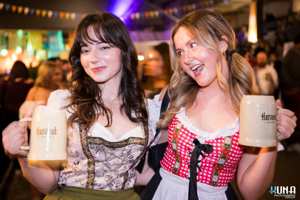 vancouver event photography - Two women in traditional German-style outfits smiling and holding up foaming Harvest Haus beer mugs at the event.
