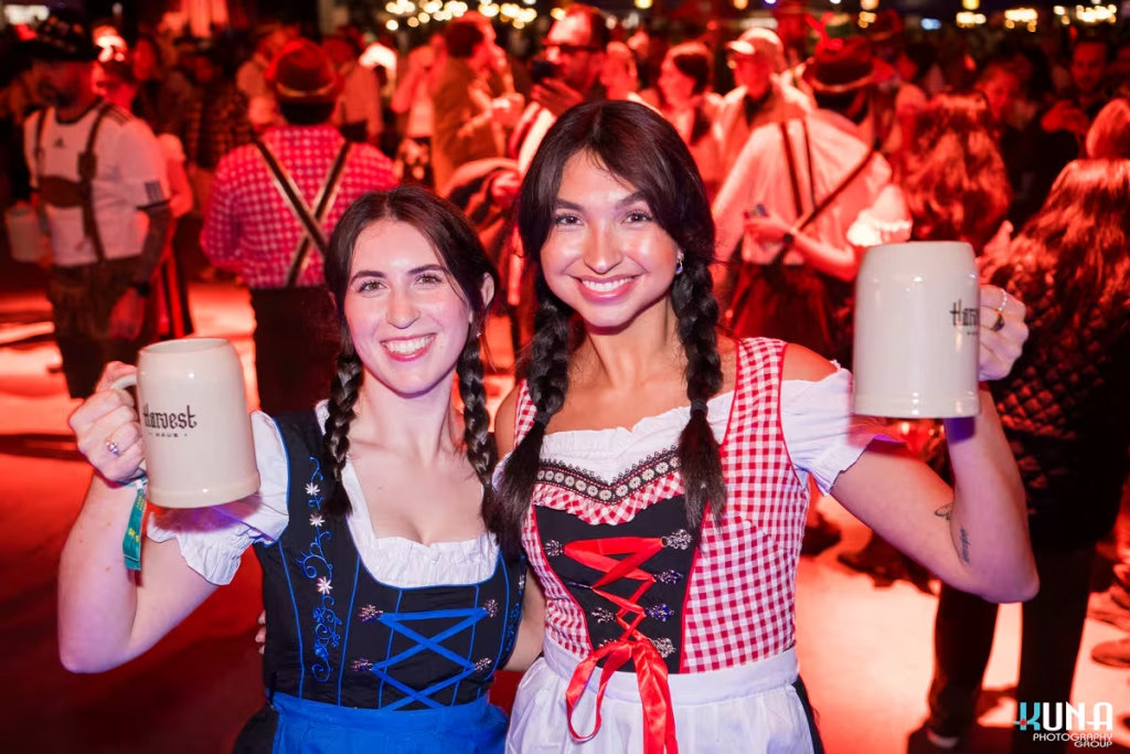 vancouver event photography - Two women in traditional dirndl dresses smiling and raising their Harvest Haus beer mugs in a festively lit hall.