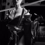 Vancouver cosplay photographer capturing a black and white photo of a female cosplayer wearing a dark, intricately textured kimono, holding a katana resting on her shoulder, and elegantly gripping a kiseru (Japanese pipe). The background features a dark, traditional Japanese shrine with strong light-and-shadow contrast.