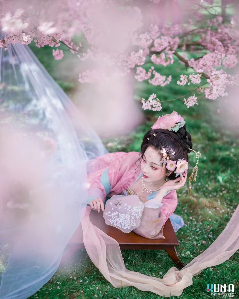 Woman in Hanfu lying playfully on a wooden desk among spring flowers.