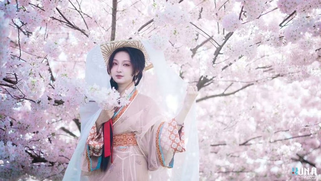 Close-up hanfu photography in Richmond BC: Model in traditional attire adjusting a veiled bamboo hat among cherry blossoms at Larry Berg Flight Path Park 2025.