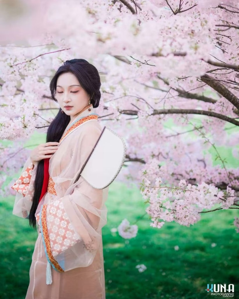 Traditional hanfu portrait in Vancouver: Subject standing beside a blooming cherry tree trunk holding a white fan at Larry Berg Flight Path Park Richmond BC 2025.