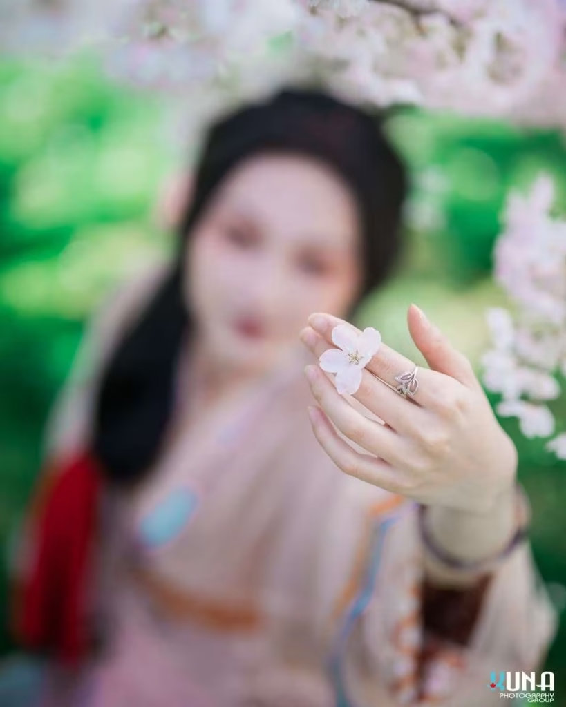 Hanfu photography bokeh detail in Metro Vancouver: Focus on a hand holding a single cherry blossom with an out-of-focus model at Larry Berg Flight Path Park 2025.