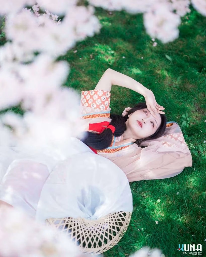 Top-down view hanfu photography in Richmond: Model lying on green grass beneath a blooming cherry canopy holding hand to forehead at Larry Berg Flight Path Park Metro Vancouver 2025.