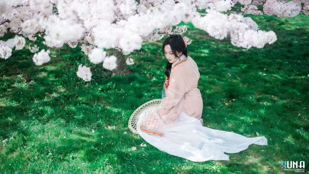 Top-down view hanfu portrait in Richmond BC: Subject seated on grass under a tunnel of cherry trees with hat beside her at Larry Berg Flight Path Park Vancouver 2025.