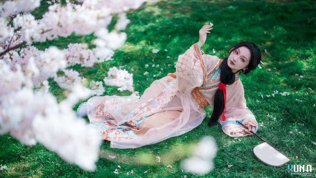 Graceful hanfu portrait in Metro Vancouver: Subject reclining on lush grass among widespread cherry groves holding a fan at Larry Berg Flight Path Park Richmond BC 2025.