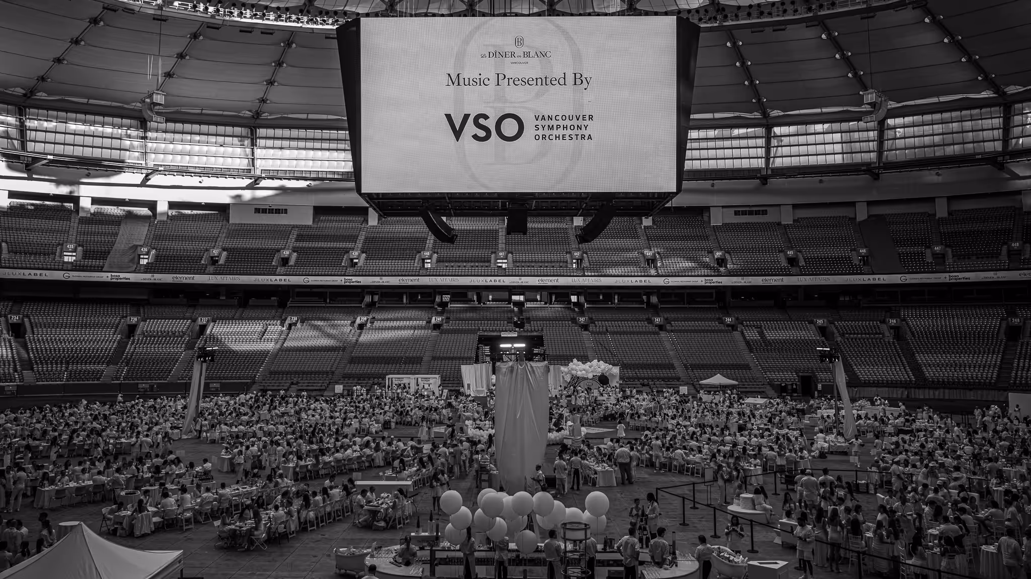 Black and white wide-angle photograph of Diner en Blanc Vancouver inside BC Place stadium with Vancouver Symphony Orchestra screen presentation, captured by Vancouver Event Photographer - Kuna Photography